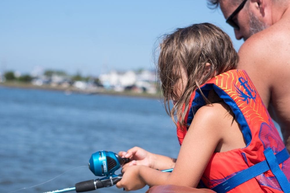 A close up of a young girl in an orange life jacket holding a blue fishing pole, her dad is behind her. Fishing is a great way to recharge your batteries as a family.
