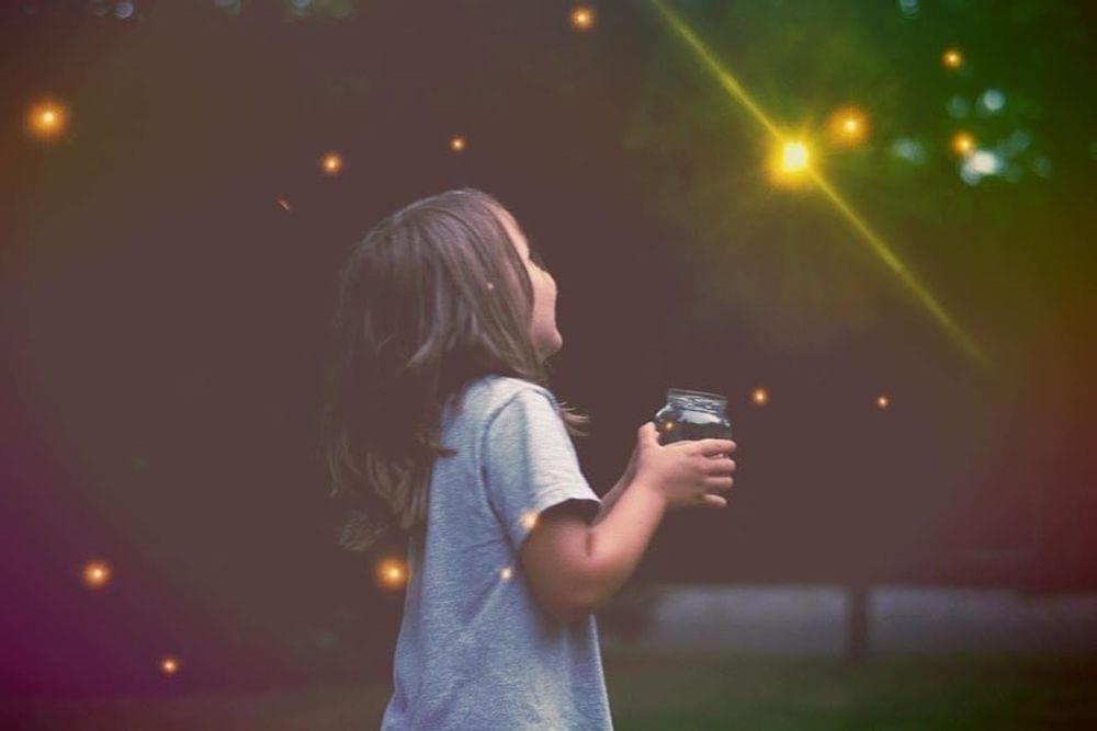 A young girl smiles up at a number of fireflies while holding a mason jar in her hand.