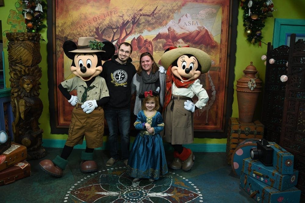 Mickey Mouse and Minnie Mouse stand on either side of two parents, while wearing safari outfits. A young firl stands in front of them.