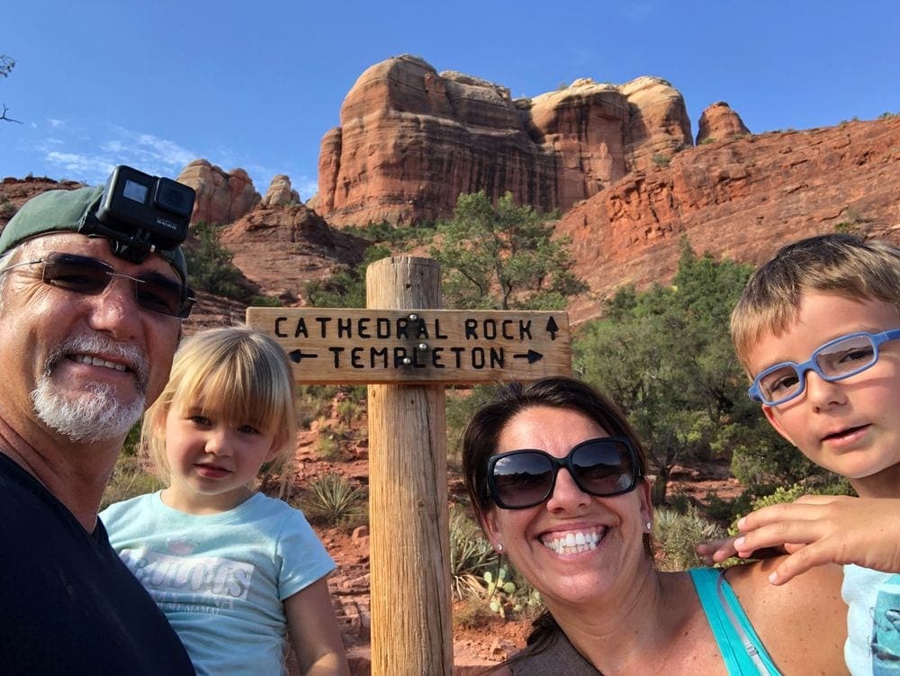 A family of four takes a selfie in front of the sign for the Cathedral Rock, pictured in the background.