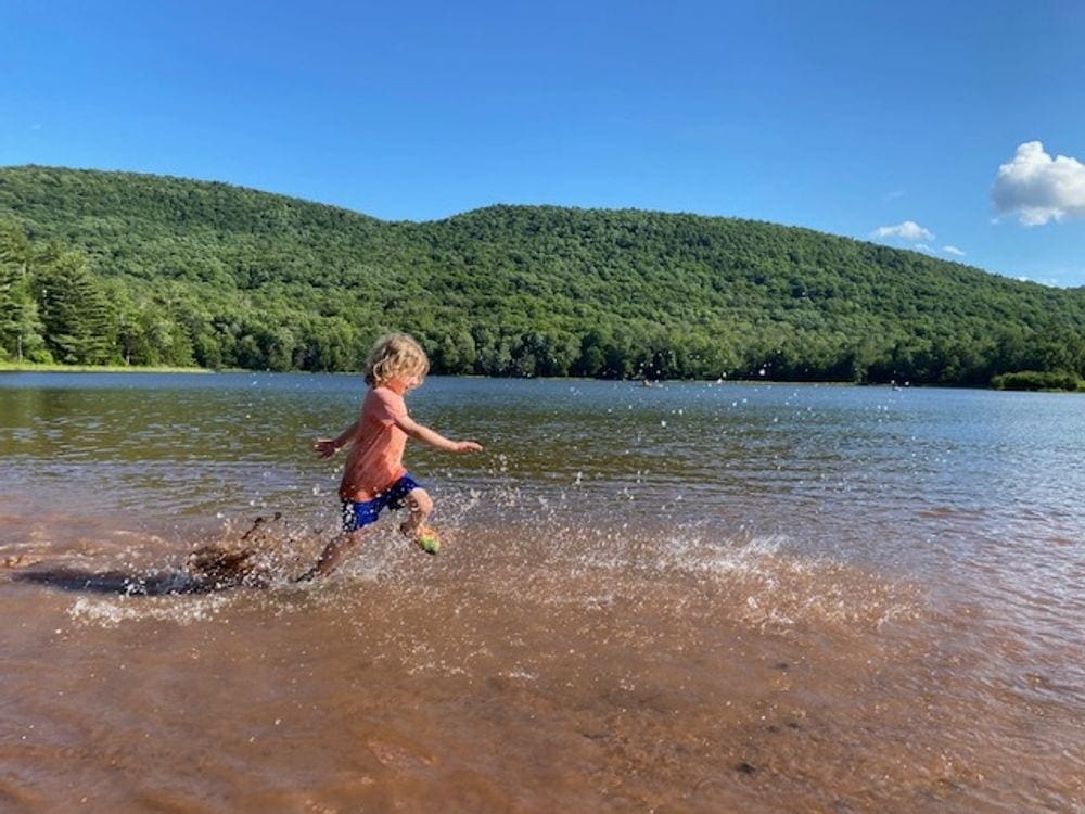 A young child wearing an orange shirt runs through shallow waters at Colgate Lake creating huge splashes around his legs.