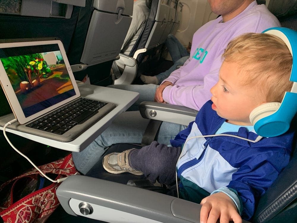 A toddler boy wearing blue headphones watches a cartoon on an iPad mid-flight.
