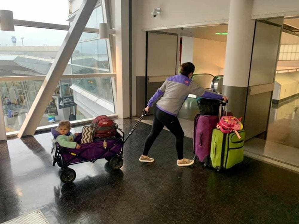 A mom pulls a toddler and luggage in a purple wagon while pushing two other bags down an airport terminal.