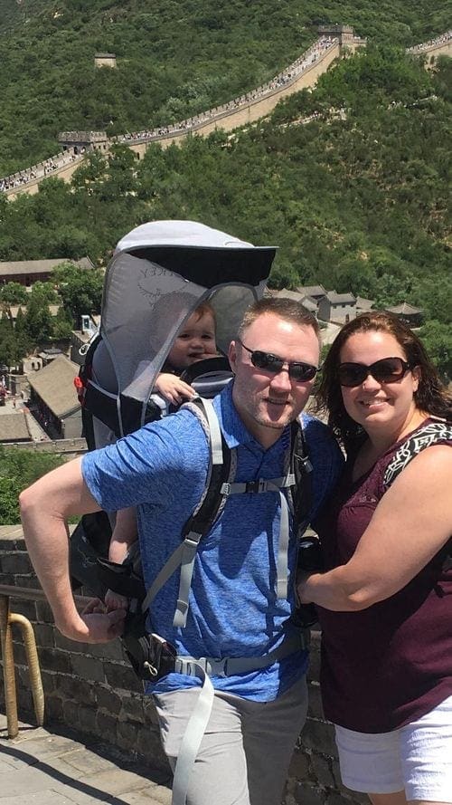 Two parents stand on the Great Wall of China, while the dad wears their infant on his back. An expansive view of the Great Wall of China is behind them.