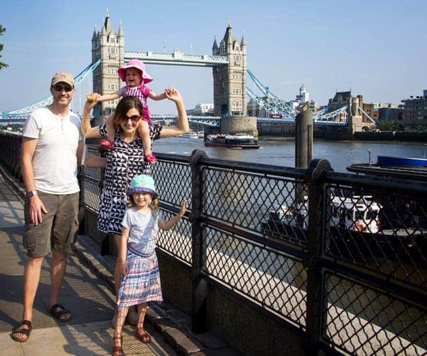 A happy family of four stands in front of the London Bridge, the youngest daughter rides on the shoulders of her mom.