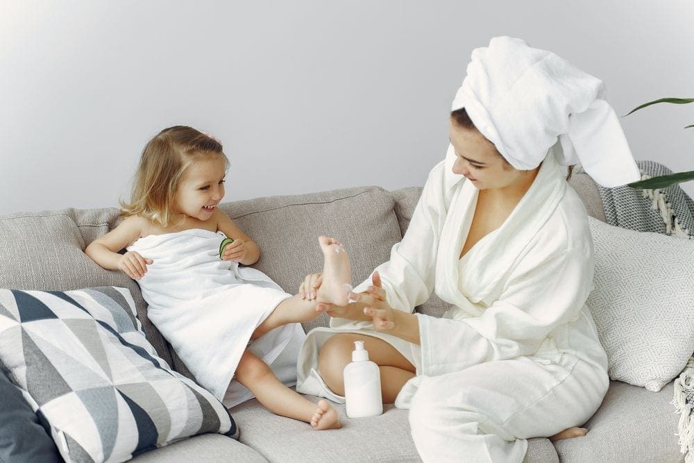 A mom wearing a robe and towel on her head rubs lotion on her young daughter's foot, also wearing a towel around her body, as they enjoy a spa day at home.