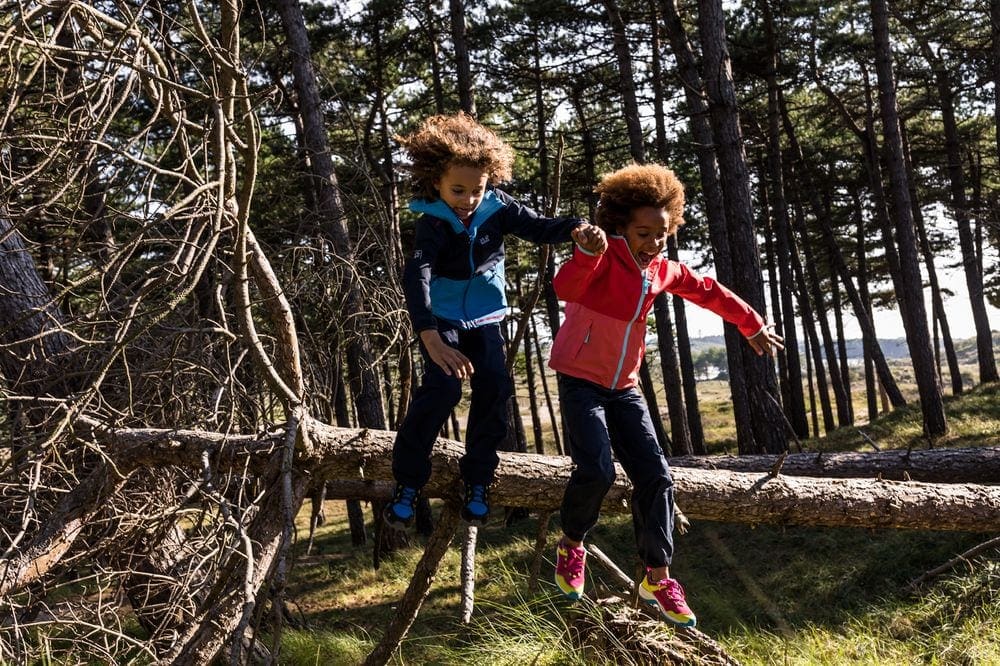 Two kids of color jump off a large log in the woods, wearing their Jack Wolfskin anti-mosquito clothing, a fantastic alternative to bug spray.