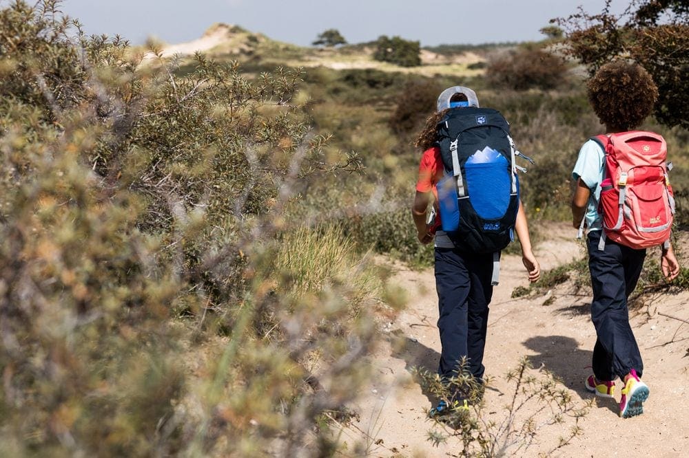 Two children of color hike on a desert trail with large packs on their backs. Both are wearing Jack Wolfskin Lakside pants for kids, which are a great alternative to bug spray.