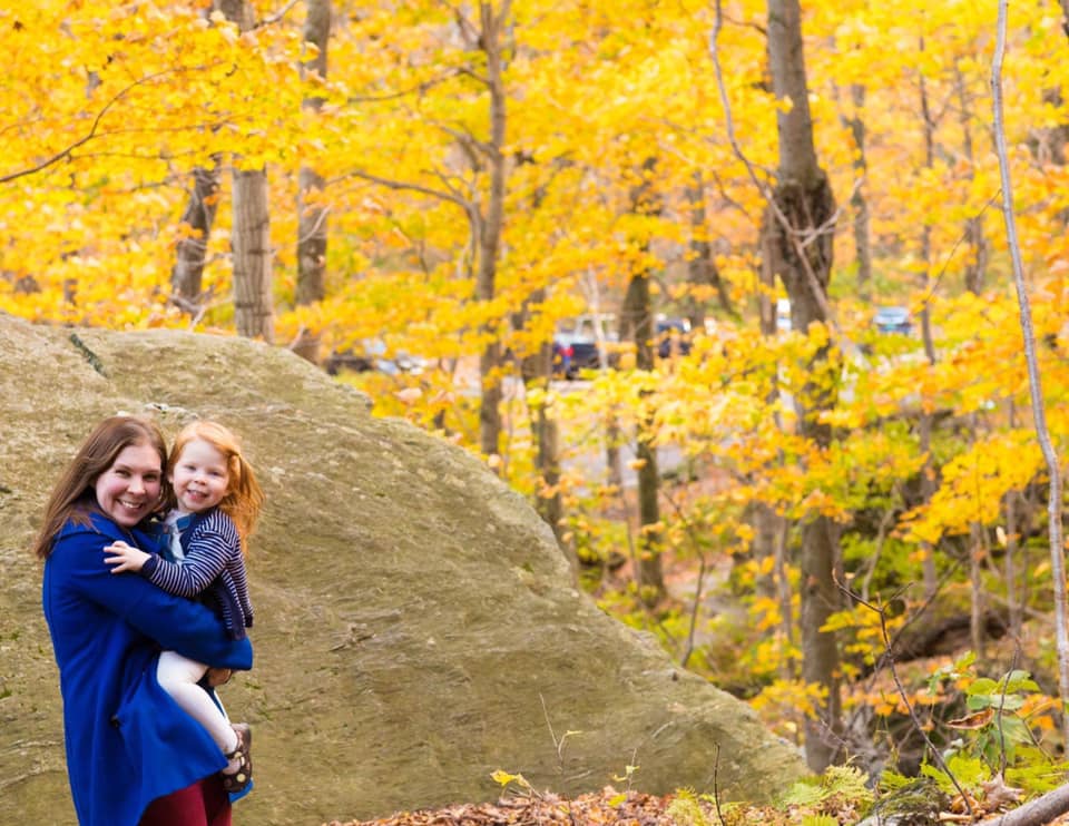 A mom hugs her young daughter in front of a large bolder, which is surrounded by a golden hues from the fall trees. Both are wearing blue coats, a striking color against the vast yellow.