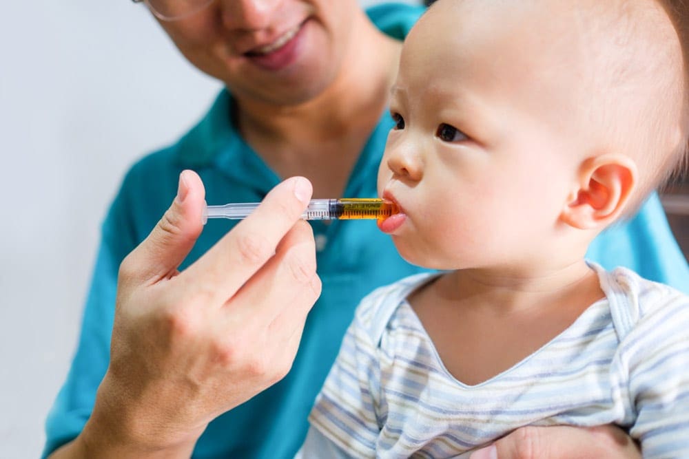 An adult administers orange liquid medication through a syringe to an infant. Kid-friendly over the counter medication is essential for any travel first aid kit for kids.