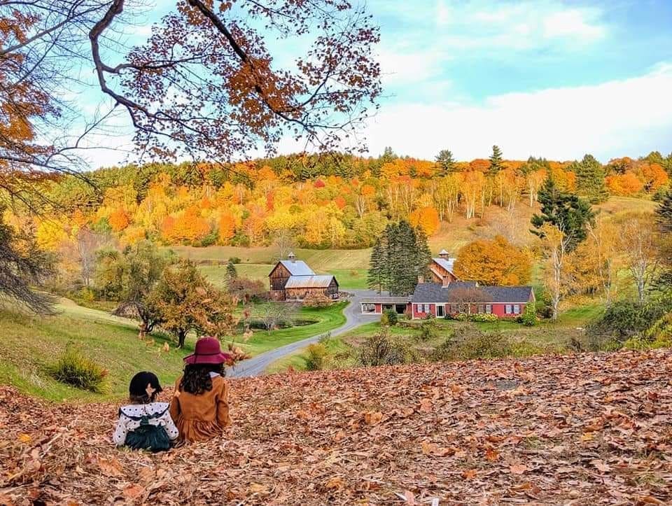 Two children sit in a vast pile of leaves looking over a small farm gilded with fall foliage in Vermont, one of the best places for fall foliage in New England.