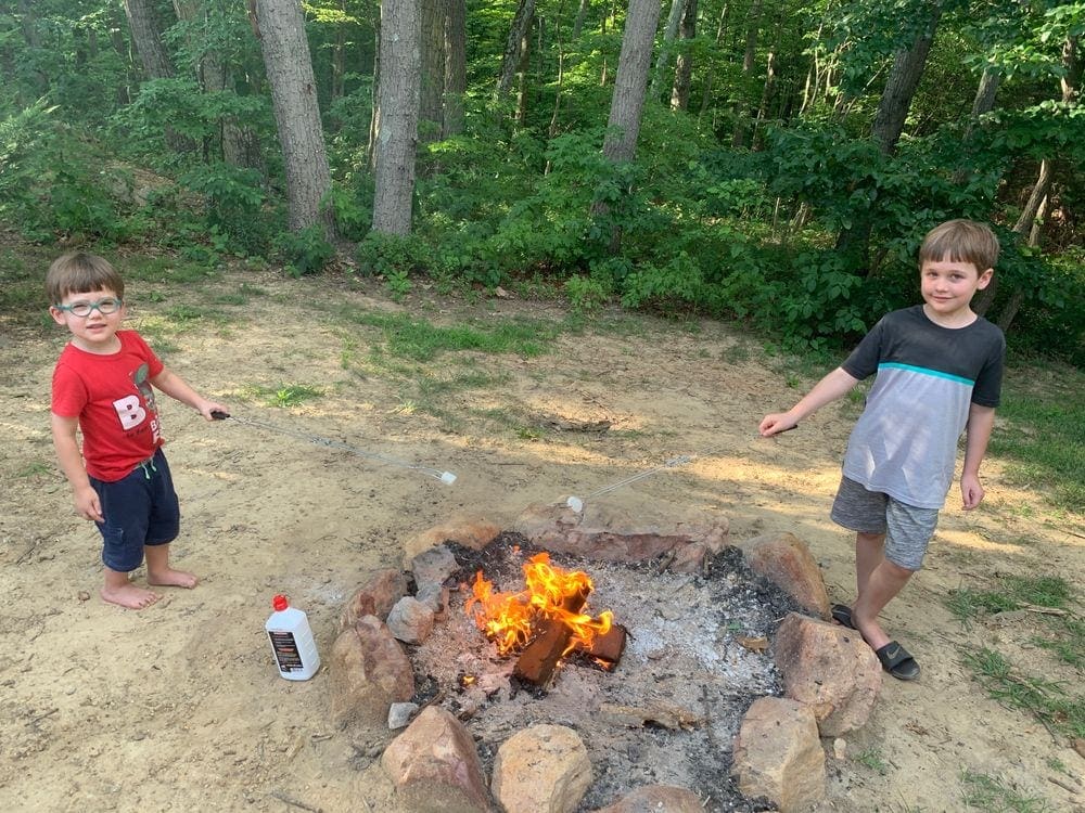 Two boys stand on opposite sides of a bonfire roasting marshmallows. Packing or researching food options beforehand will help you feel more prepared when you travel with kids during. COVID-19.