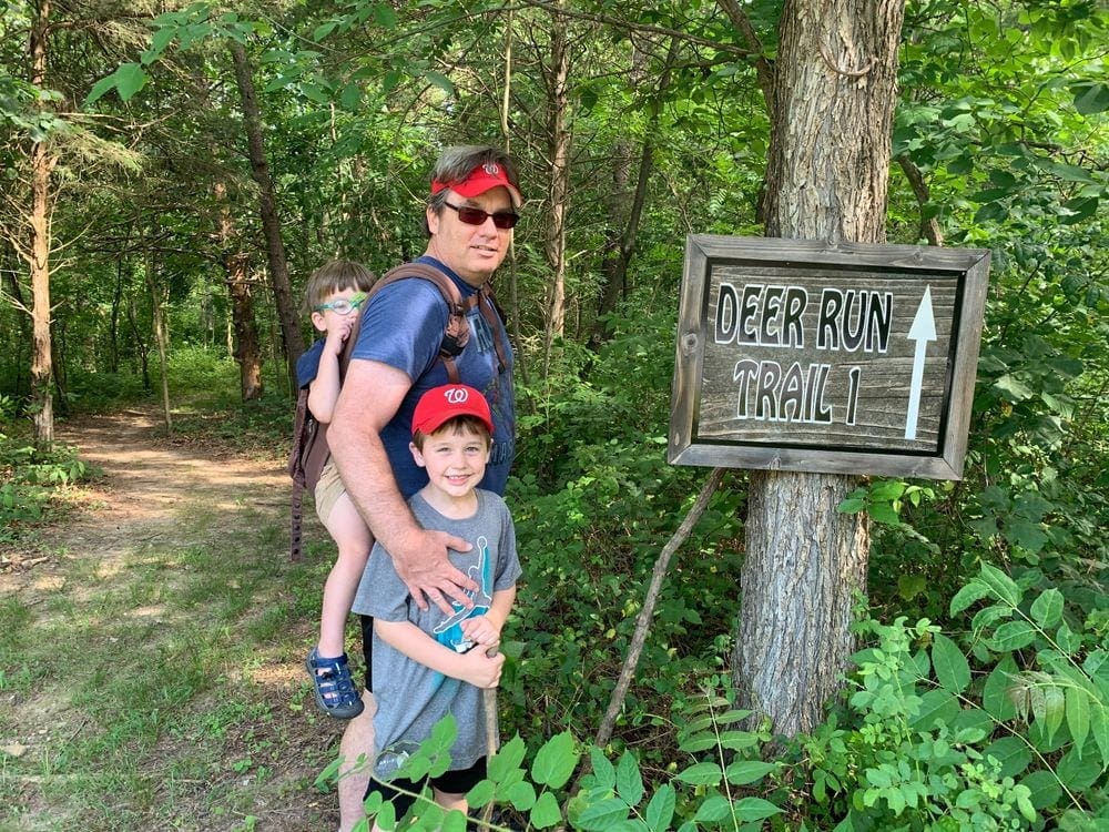 A dad stands need a trail sign that reads "Deer Run Trail 1", while his older son stands in front of him and the younger son sits in a back carrier on his back.