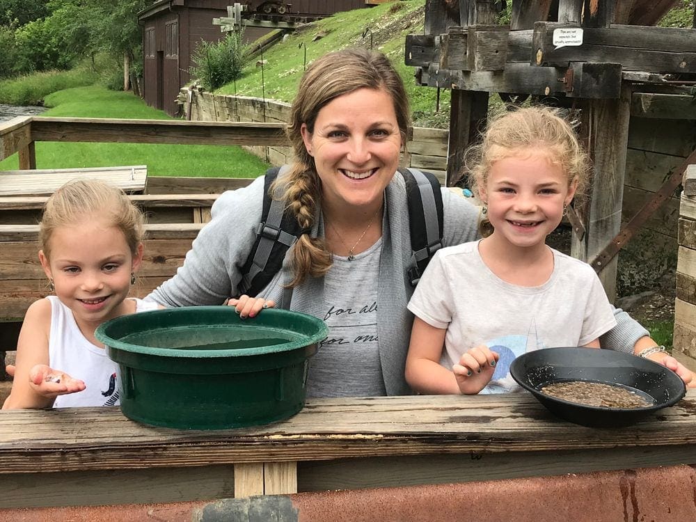 A mom and her two girls sit smiling with gold panning tools in front of them. Creating education experiences around the world is one of the benefits of traveling with kids.