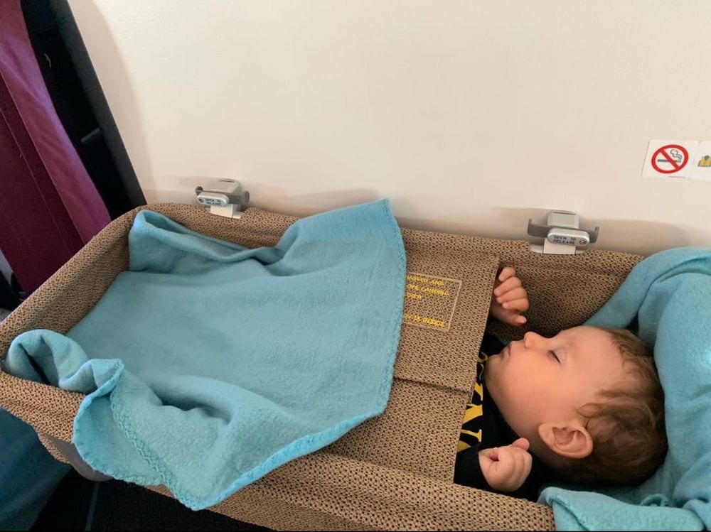 An infant boy snuggles with a blue blanket while sleeping in an on-board bassinet mid-flight.