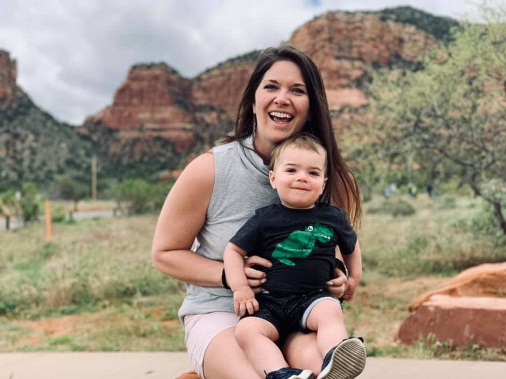 A smiling mother holds her infant with an expansive view of the inconic Red Rocks of Sedona behind them. Exploring Red Rock State Park is one of the best things to do in Sedona for families.