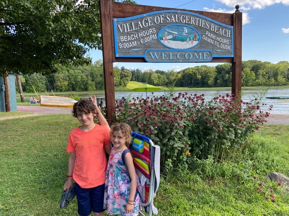 Two kids stand with goofy poses in front of the official sign for the Village of Saugerties Beach.