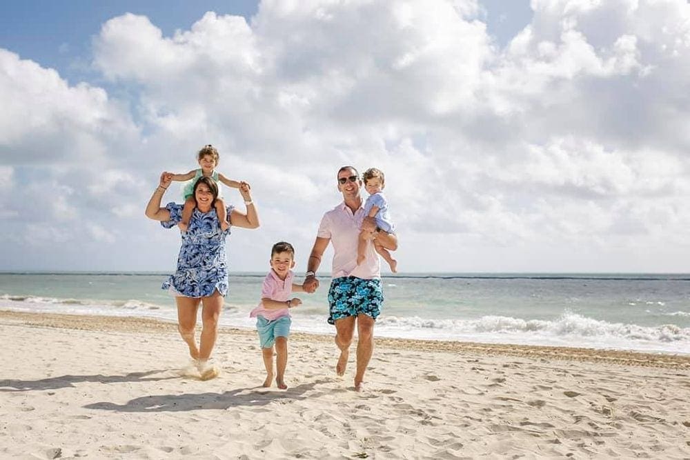 A family of five walks along a beach in Cancun. The mom holds a young girl on her shoulders, while the dad holds another small child and the hand of a third child.