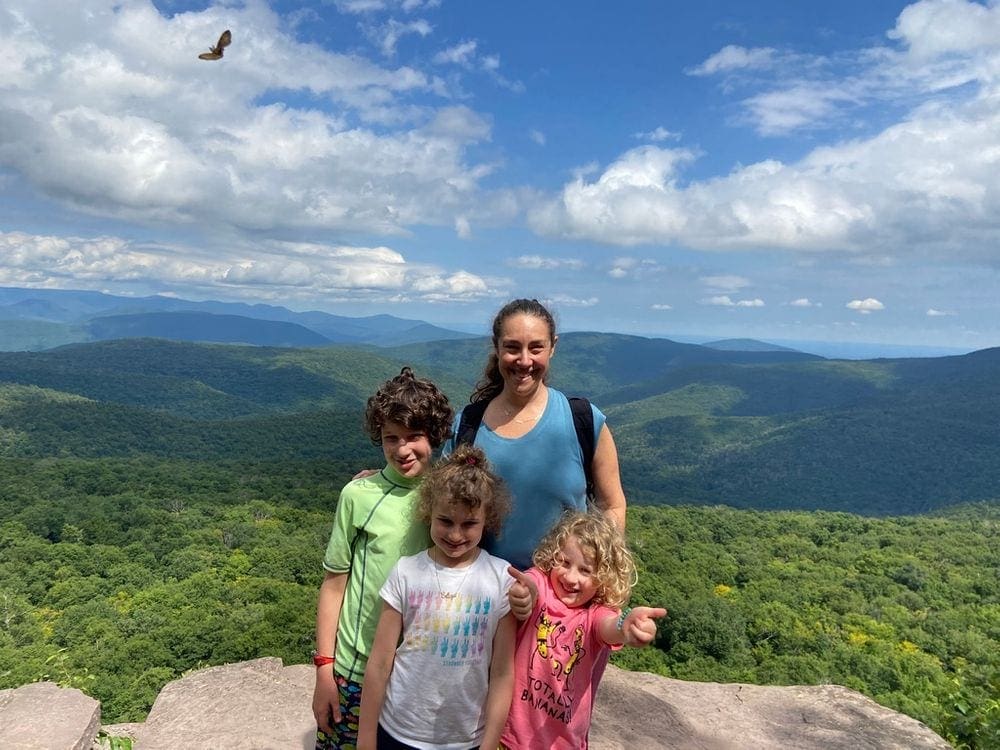 A mom and her three kids stand happily on a giant ledge within the Catskill Mountains at the top of the Giant Ledge Trail, one of the most kid-friendly hikes in the Hudson Valley.