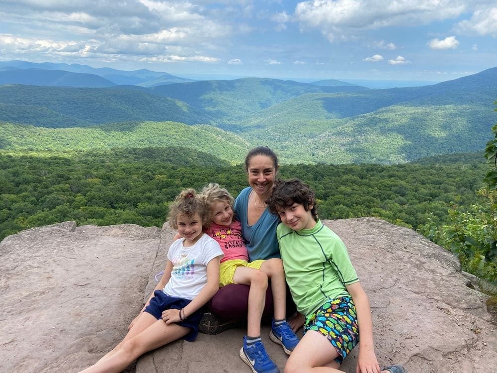 A mother holds two children on her lap, while a third sits closely to her, on a rock ledge overlooking the lush Catskill Mountains.