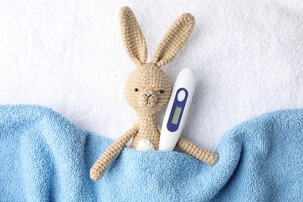 A small stuffed bunny and a thermometer are tucked into a blue blanket. A thermometer is a must for any travel first aid kit for kids.