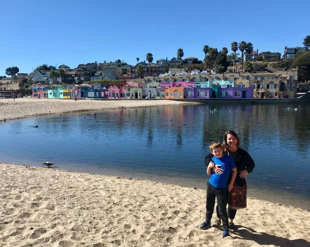 A mom put her arm around her older son as they stand on the beach, behind them glistening water lies in front of a colorful town on the California coast.
