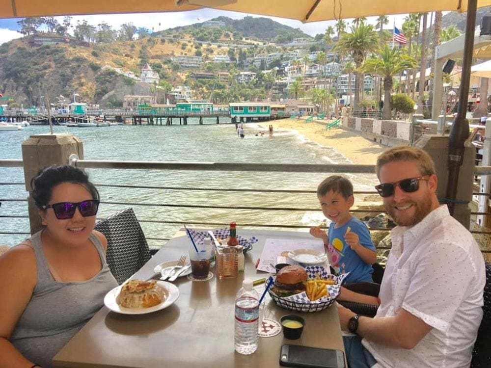 A mom, dad, and their young son enjoys lunch while a colorful array of homes and coastline dots the backfound of one of the most charming towns to visit with kids, Santa Catalina Island.