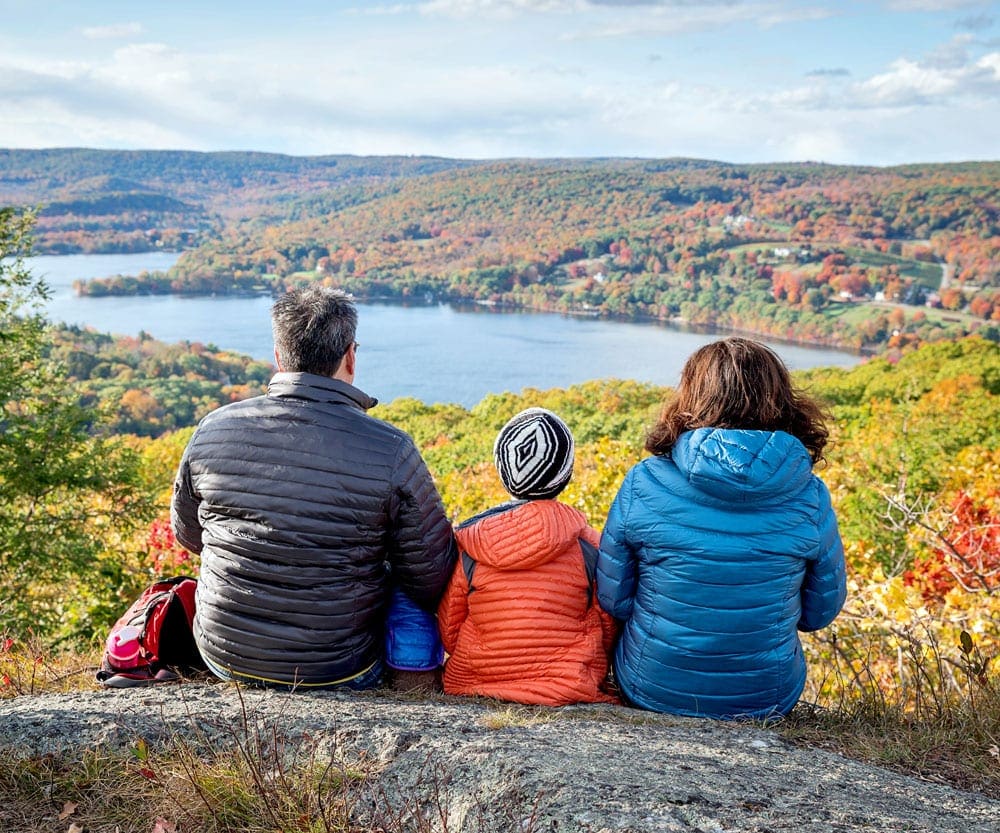 A family of three sits upon a large boulder overlooking a colorful autumn display in New England, a beautiful destination is one of the best ways to add romance on a family road trip.