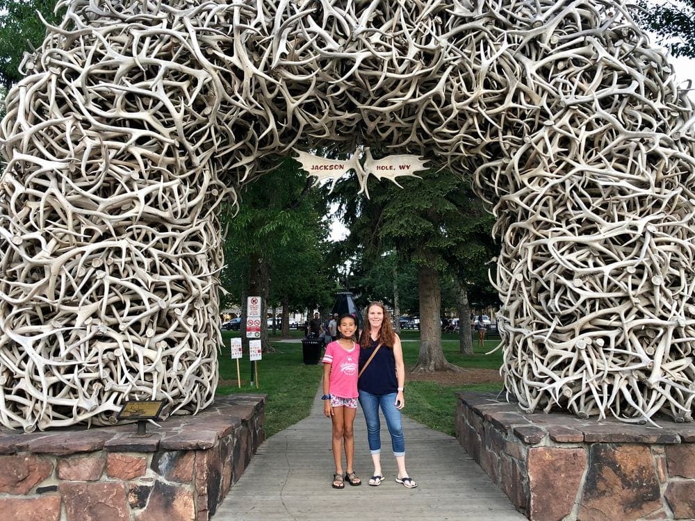 A mom and her pre-teen daughter stand in front of Jackson's iconic entrance created with fallen antlers. Jackson, Wyoming is one of the most charming towns to visit with kids.