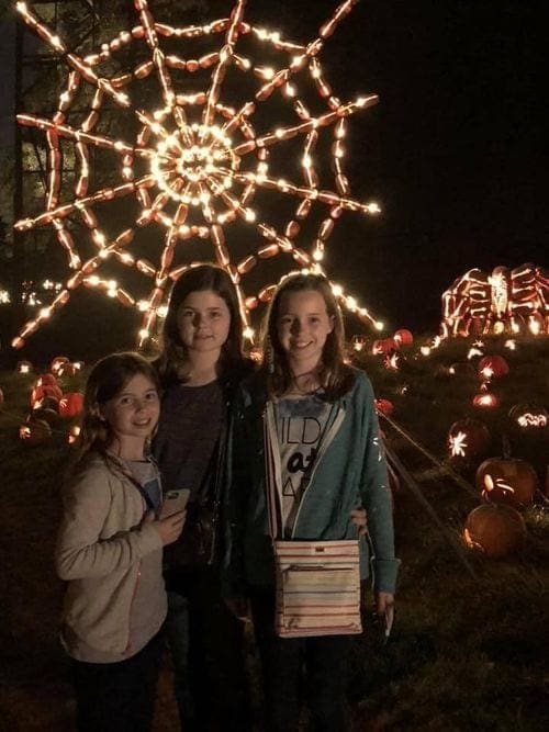 Three young girls stand in front of a lit up spider web at the Great Jackolantern Blaze in Sleepy Hollow.