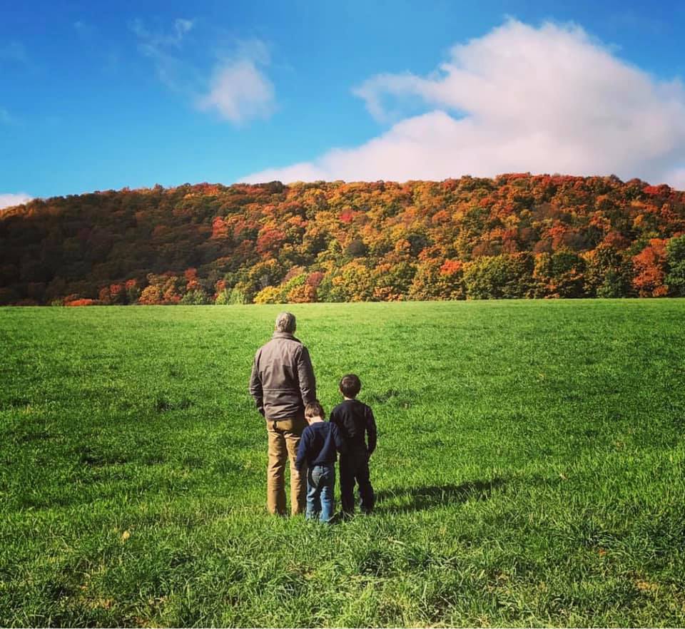 A father stands with his two sons gazing upon a moutain awash in fall hues of orange, yellow, and auburn. Kids will love taking a fall foliage train ride to mark the season.