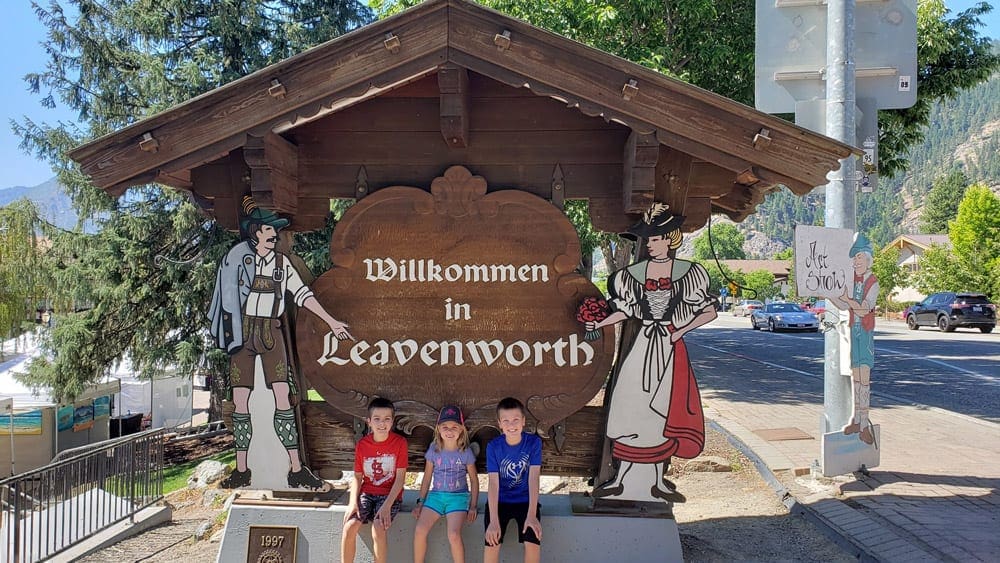 Three kids sit in front of a sign reading "Wilkommen in Leavenworth", which is one of the most charming towns to visit with kids!