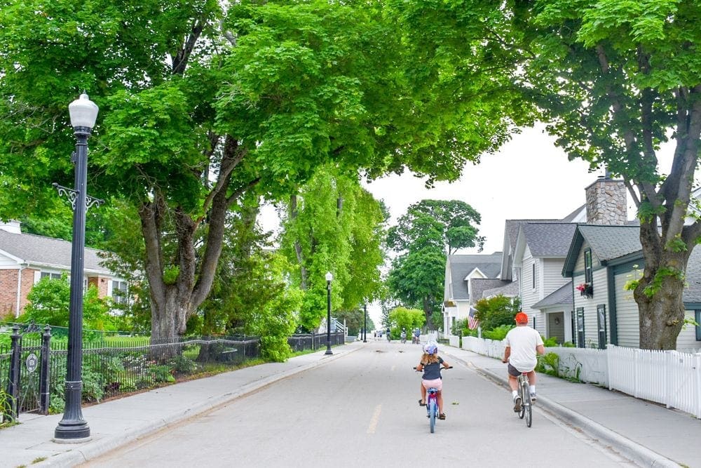 A young girl and her father bike along a charming, tree-lined streen on Mackinac Island, Michigan, a great stop for a Midwest road trip with kids.