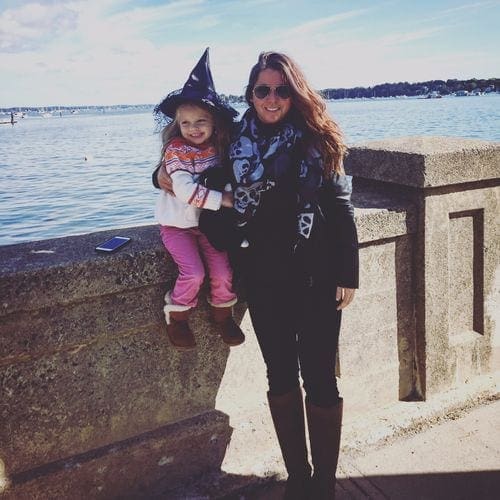 A mom stands holding her young daughter, who is wearing a witch hat, along the waterfront in Salem.