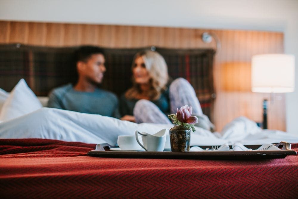 A tray of coffee and treats sits at the edge of a bed, while two adults sit talking in the background.