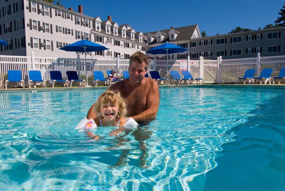 A dad holds his toddler daughter by the waist while she tries to swim in a stunning outdoor pool at The Nonantum Resort, one of the best Maine hotels for families.