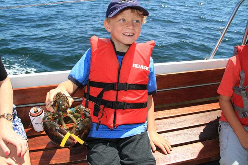 A young boy wearing an orange life vest holds a lobster while on a boating excursion.