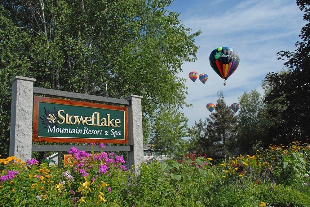 The sign for Stoweflake Mountain Resort & Spa emerges from an array of colorful flowers with several hot air balloons in the air behind it.
