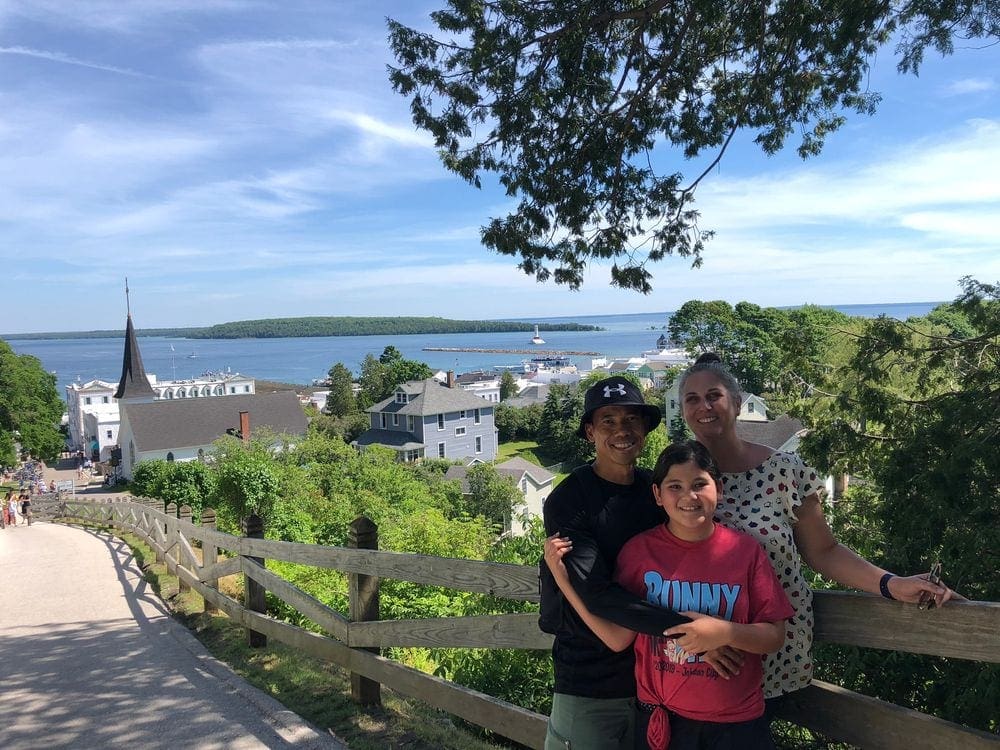 Parents put their arms around their pre-teen son, with an expansive view of Mackinac Island behind them, featuring charming buildings and greenery.