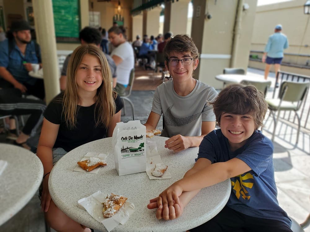 Three kids sit around a table at Cafe du Monde enjoying their beignets.