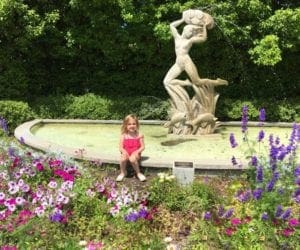 A young girl wearing pink sits next to a fountain surrounded by blooming flowers in City Park.
