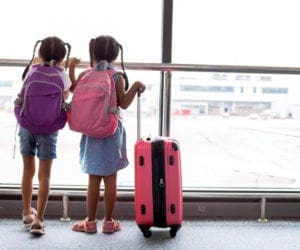 Two girls wearing backpacks, one purple and one pink, respecitve, stand next to a pink carryy-on while they look out a large window onto planes at the airport.