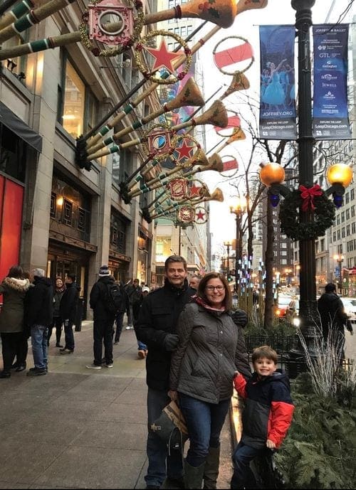 A family of three stands smiling on a street in Chicago under a number of holiday decorations.