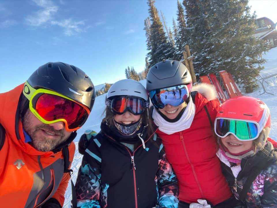 A family of four wearing large snowcoats, helmets, and googles huddles together smiling.