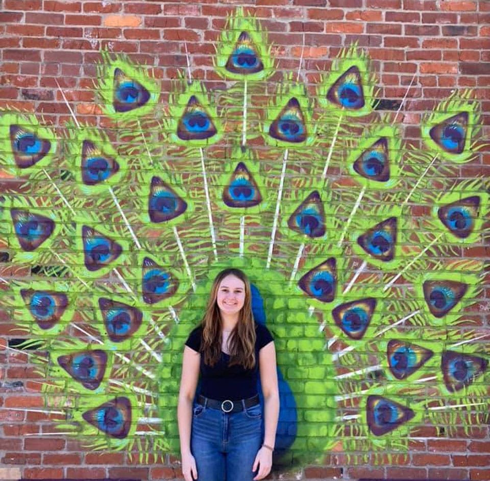 A woman stands in the center of street art, with peacock feathers fanning behind her in hues of green and blue.
