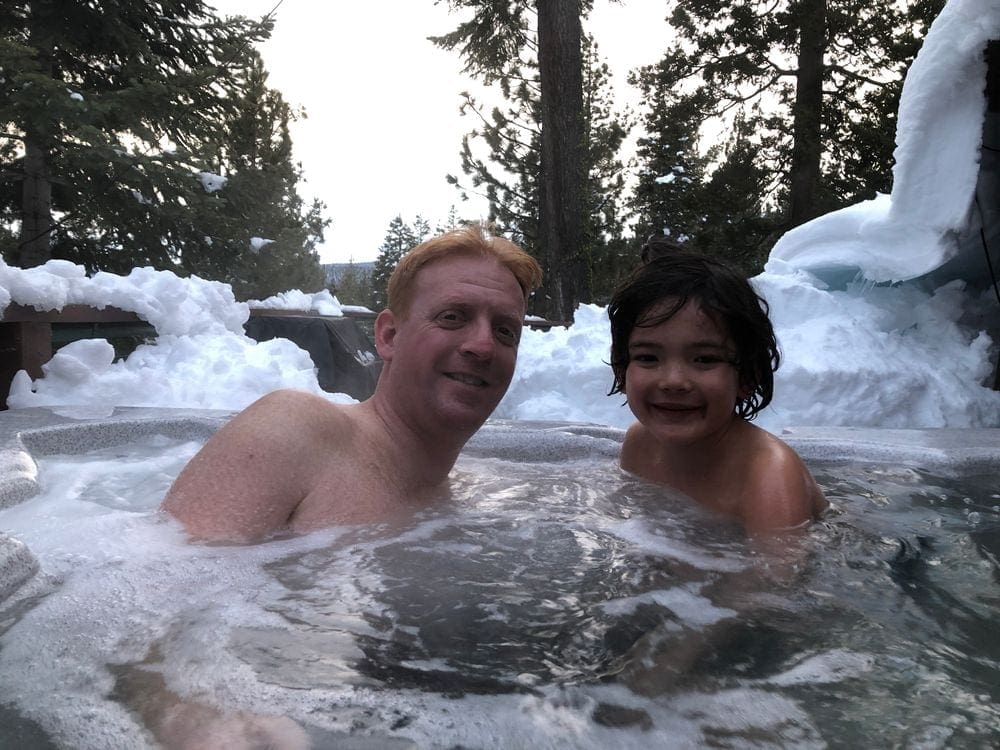 A man and his young son enjoy a hottub surrounded by snow.