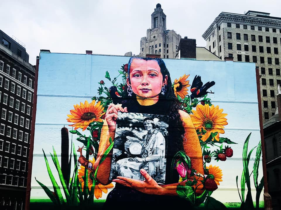 In Rhode Island, a street mural of an indigenous woman holding a black and white photo of another indigenous woman, with sunflowers in the background.