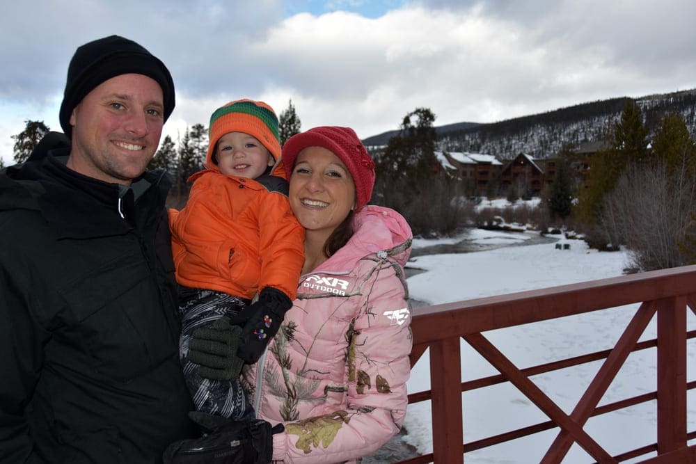 A family of three wearing snow gear stands on a bridge in Keystone, one of the best Colorado ski resorts for families.