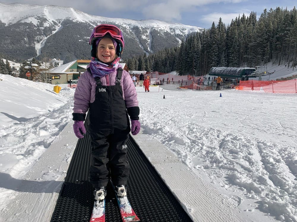 A young girl wearing snow gear stands on skis smiling while learning to ski at Copper Mountain.