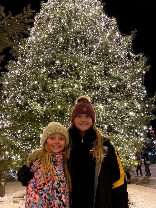 Two tween girls stand in front of a lit-up Christmas tree in Breckenridge, Colorado, one of most magical Christmas towns for families.
